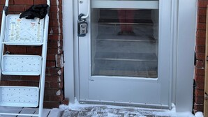 Een witte deur met een ruit, een metalen trapzetter met een zwarte tas, en een bakstenen muur.