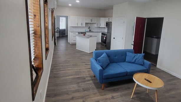 Een moderne woonkamer met een blauwe bank en een ronde salontafel, een keuken met witte kasten en een gang die naar een andere kamer leidt.