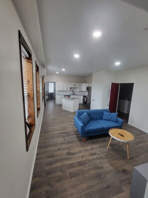 A modern living room with a blue sofa and a round coffee table, a kitchen with white cabinets, and a hallway leading to another room. - Modern Mountain Retreat Apt-3-3| High-End Finishes| Easy Highway Access (Windber)