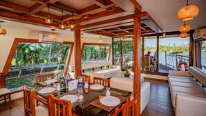 A dining area with wooden furniture, large windows, and a view of the water. - Riverine Kadavil Houseboat (Alappuzha)
