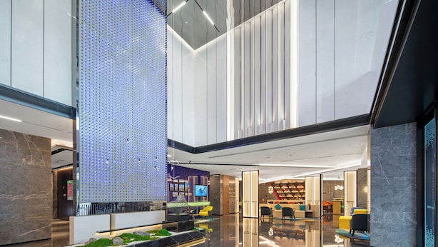 A modern lobby with a high ceiling, a seating area with yellow chairs, a reception desk, and a large digital display.