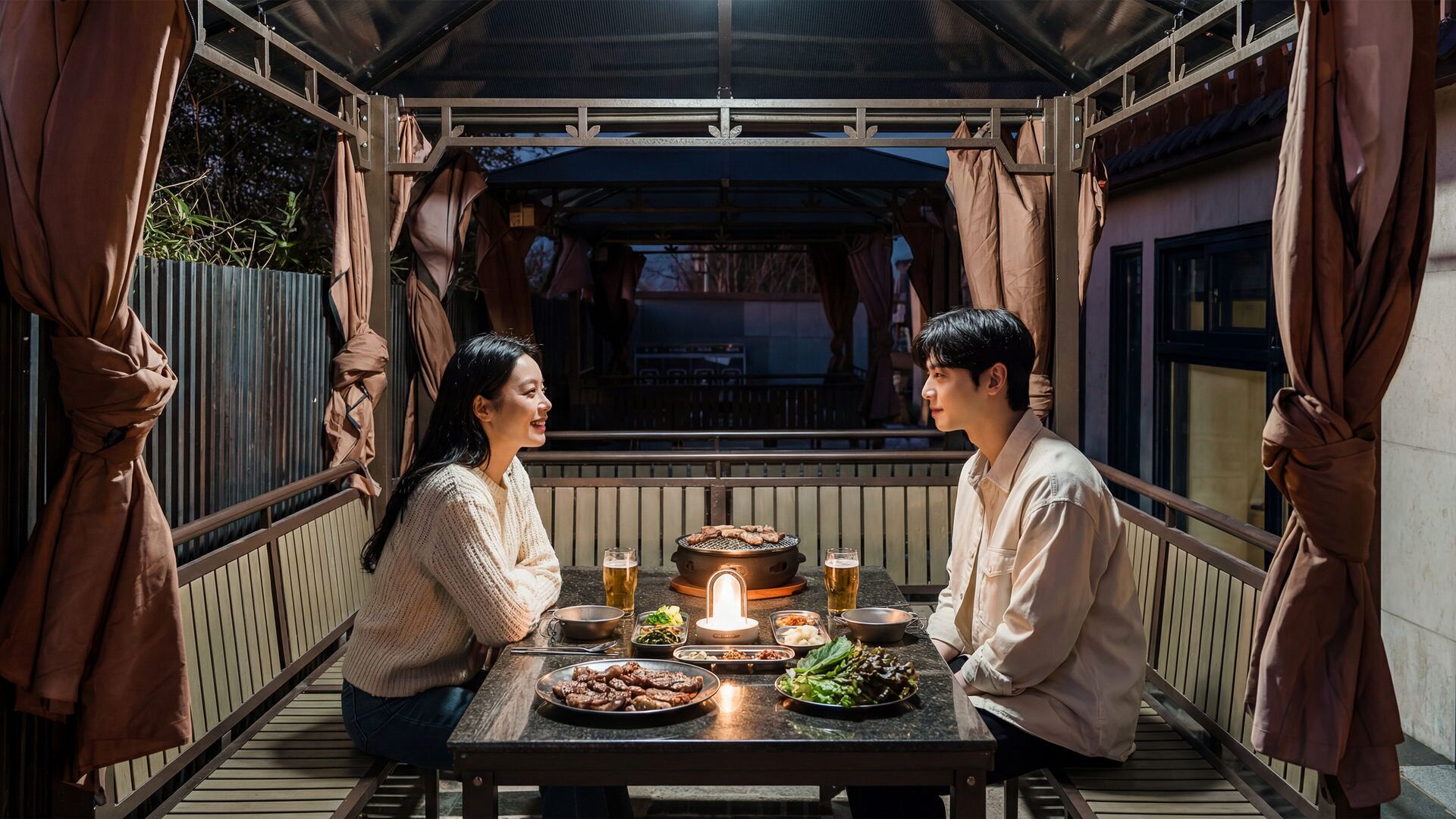 A couple dining at an outdoor table with a grill, food, and drinks.