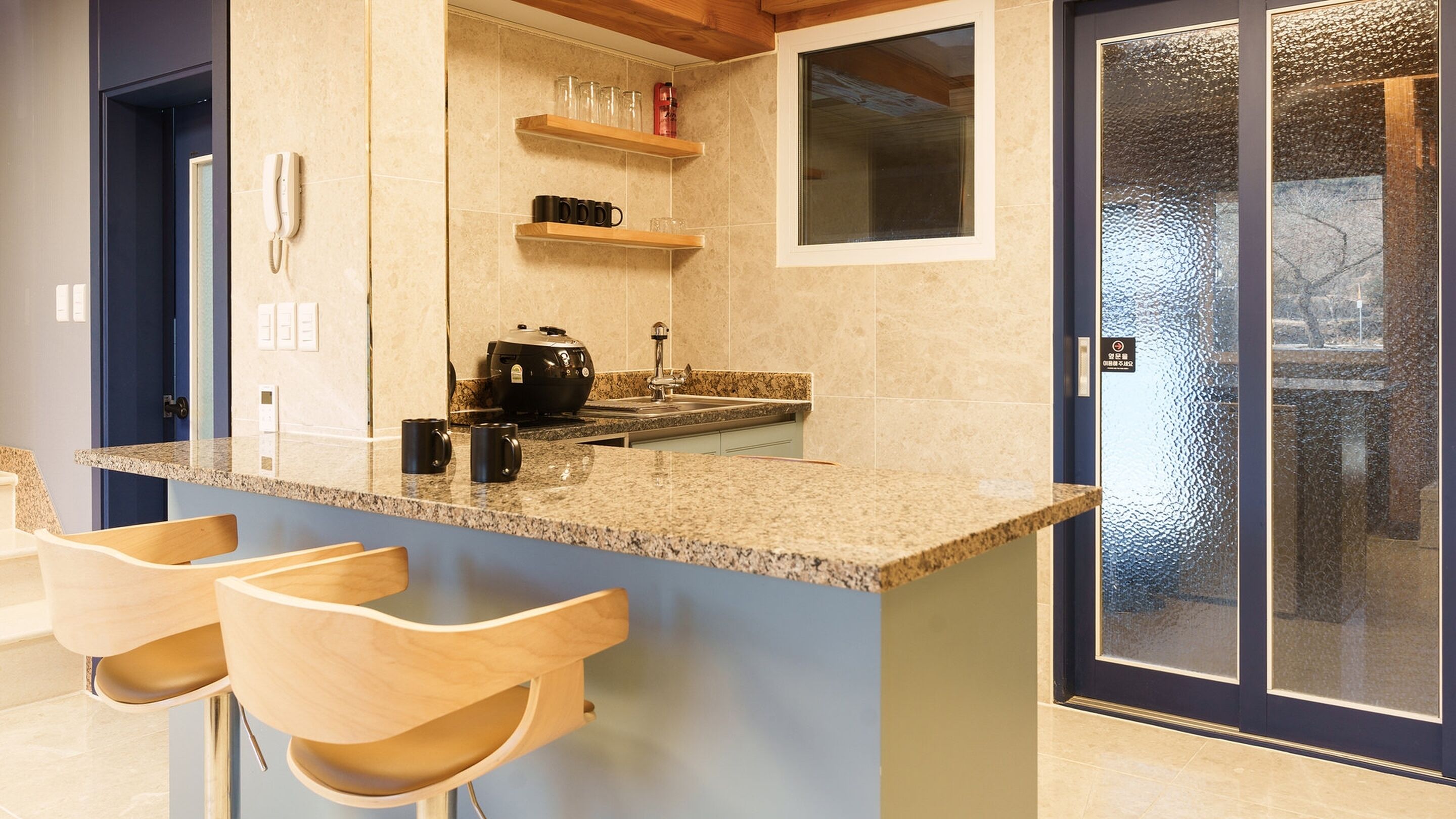 A kitchen with granite countertops, a coffee maker, and a built-in oven. There are three wooden bar stools and a double door with frosted glass panels.