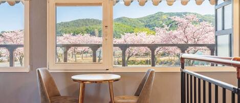 A balcony with two chairs and a small table, overlooking cherry blossoms and a hilly landscape.