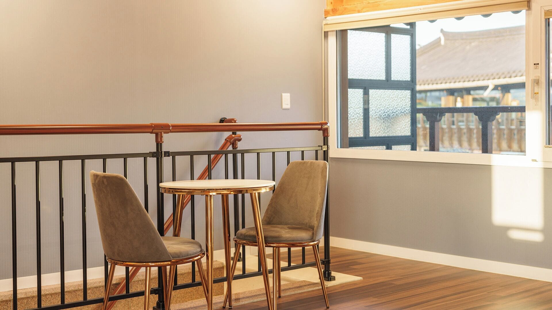 Een balkon met twee stoelen en een kleintje tafeltje, een houten leuning en uitzicht op een gebouw door een raam.