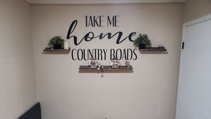 Une pièce avec une pancarte fixée au mur sur laquelle on peut lire « RAMENEZ-MOI DANS MON PAYS, OBLIGATIONS NATIONALES », un canapé noir, une petite table sur laquelle se trouve une boîte décorative et un porte-chaussures.