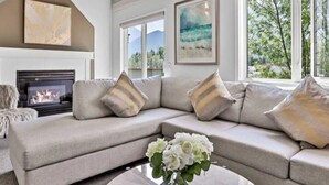 Ein modernes Wohnzimmer mit einem beigefarbenen Ecksofa, einem Kamin und einem großen Fenster mit Blick auf Bäume und Berge.