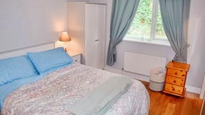 Um quarto com cama, mesa de cabeceira, luminária, guarda-roupa, janela com cortinas e cômoda de madeira.
