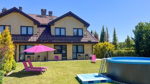 Una casa de dos pisos con piscina, un patio con sillas y un paraguas, y un césped bien cuidado.