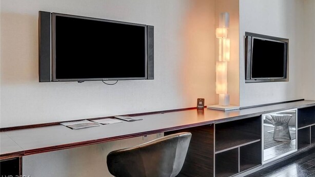 A modern hotel room with a large desk, two wall-mounted TVs, a chair, and a decorative light fixture. - Stunning Strip-view Studio w/ Balcony Access 35308 (Las Vegas)