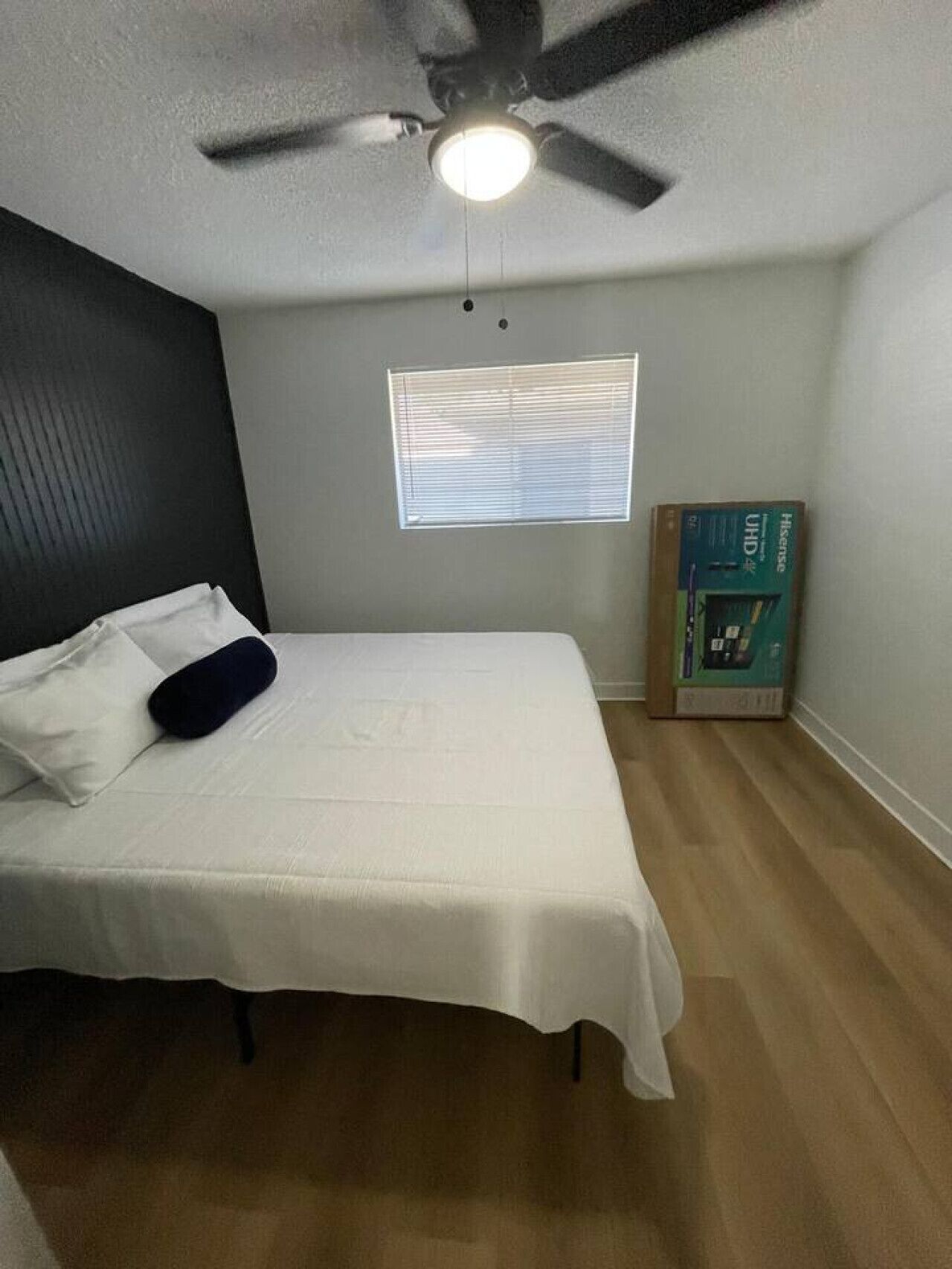 A bedroom with a bed, a ceiling fan, a window with blinds, and a box on the floor.