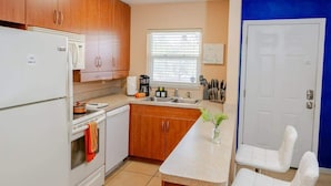 Una cocina con armarios de madera, un refrigerador blanco, una cafetera y un lavamanos. Hay una ventana con persianas y una puerta con una pared azul de fondo.
