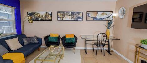 A modern living room with a blue sofa, two black chairs with green cushions, a glass coffee table, and a dining table with chairs.