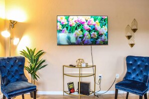 Een kamer met twee blauwe fauteuils, een aan de muur bevestigde televisie, een tafeltje met decoratie en een plant.