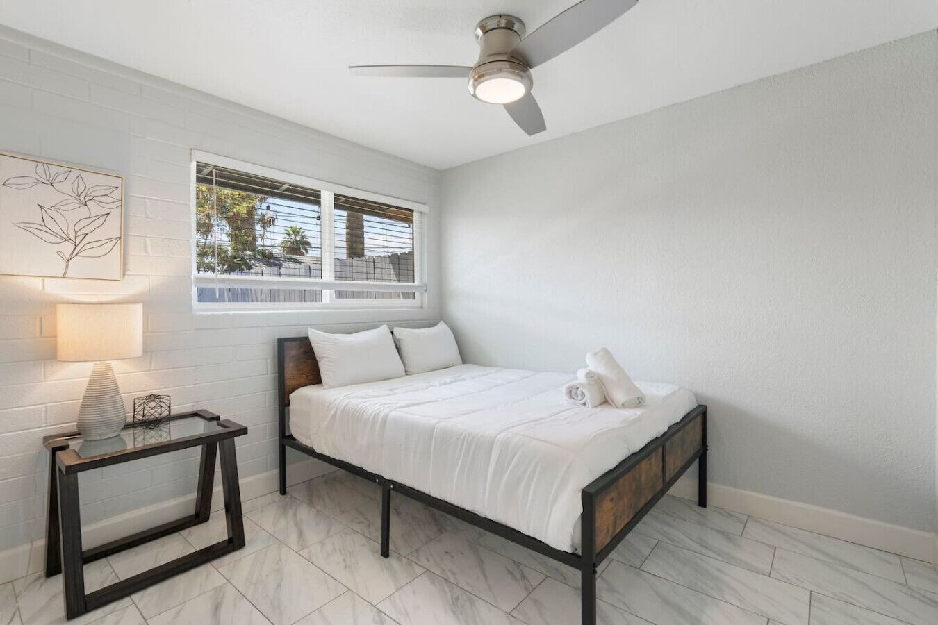 A bedroom with a bed, a nightstand, a ceiling fan, a window, and a wall-mounted artwork.