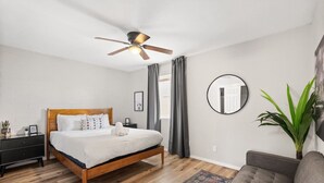A bedroom with a bed, a ceiling fan, a round mirror, a gray sofa, and a potted plant. - Fashion Square Apt3 (Scottsdale)