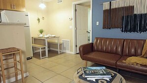 A living room with a brown leather sofa, a wooden coffee table with magazines, a wooden stool, a kitchen area with a refrigerator, and a dining table with chairs. - Calm Japandi Retreat With Patio, Near Downtown (Sarasota)