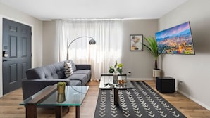 Ein modernes Wohnzimmer mit grauem Sofa, einem Glastisch, einem schwarzen Teppich, einem Fernseher und einem Stadtbild.
