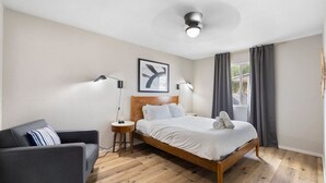 Ein modernes Hotelzimmer mit einem hölzernen Bett, einer grauen Couch, einem kleinen Beistelltisch, einem Deckenventilator und einem gerahmten Kunstwerk an der Wand.