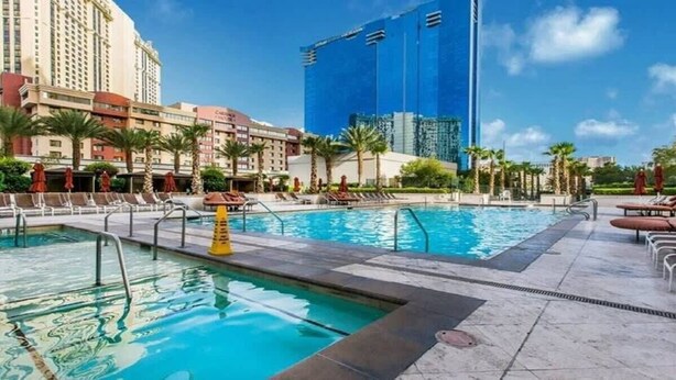 A hotel pool area with multiple pools, palm trees, and a large blue building in the background. - Free Valet Parking MGM Studio 1911 (Las Vegas)