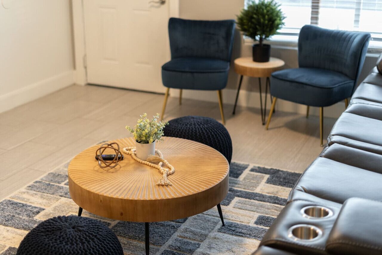 Una sala de estar moderna con una mesa de centro redonda de madera, dos sillones azules, una mesita auxiliar y una planta en maceta.