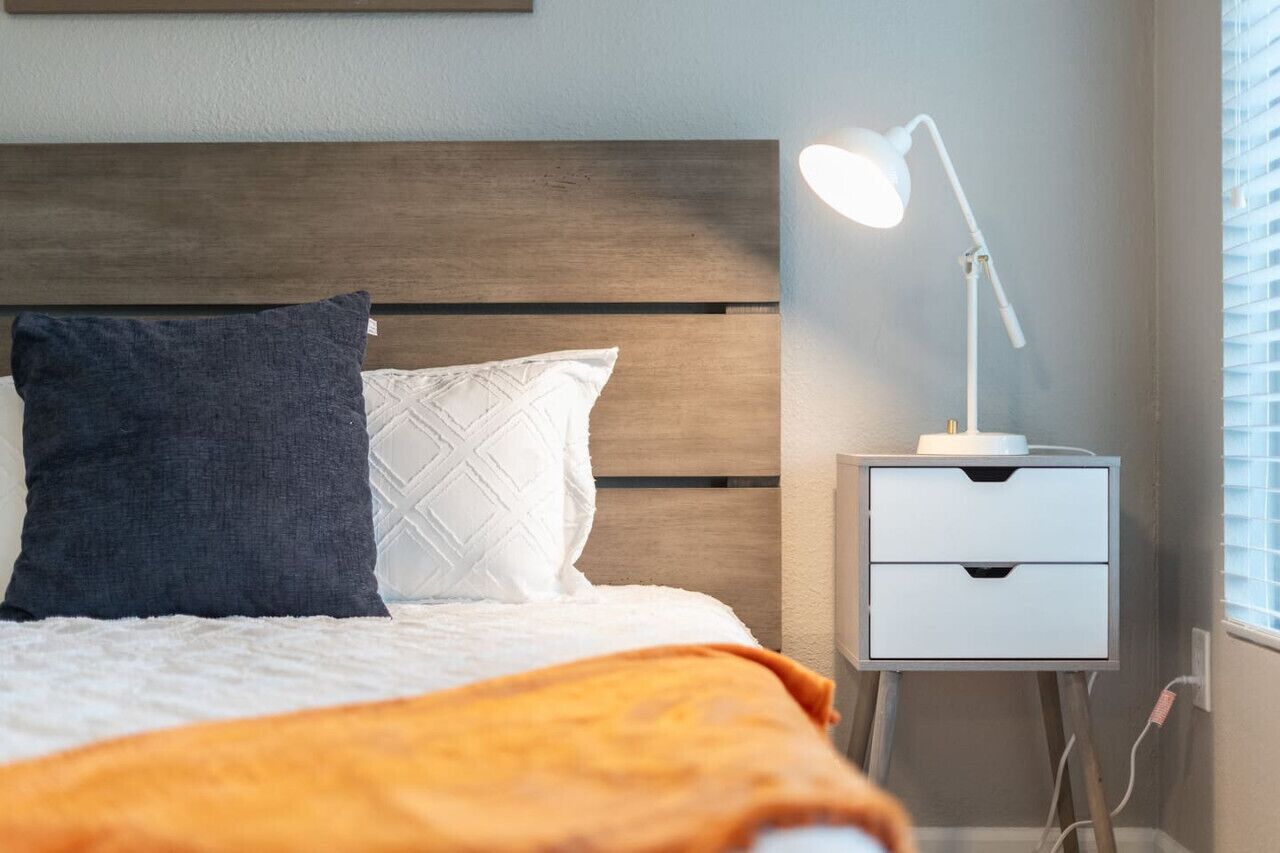 Una cama bien hecha con un almohadón blanco y azul oscuro, una mesita de noche blanca con cajones y una lámpara de escritorio blanca.