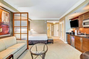 A hotel room with a bed, a sofa, a glass table, a kitchen area with a microwave, and a TV. - MGM Studio Sphere View (Las Vegas)