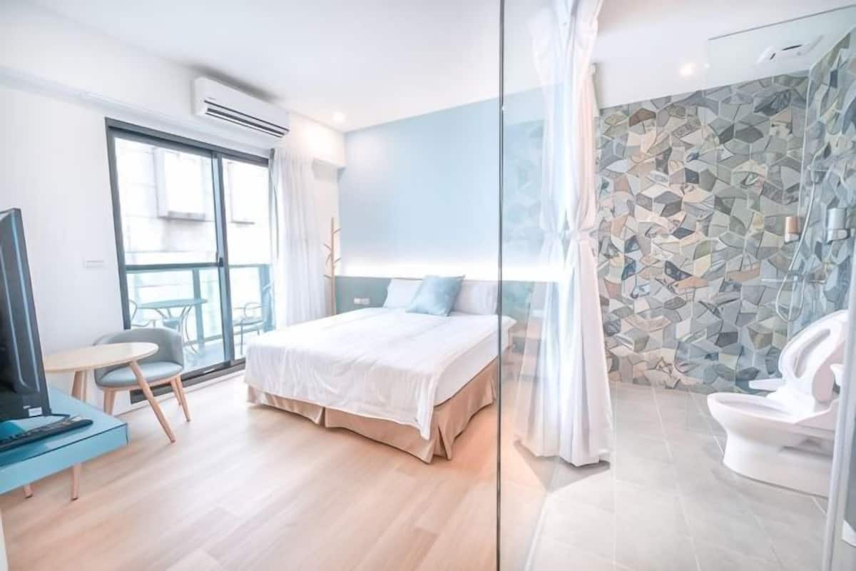 Ein modernes Hotelzimmer mit Bett, kleinem Schreibtisch, Fernseher und einer Duschkabine aus Glas. Durch die Glastür bietet es einen Ausblick.