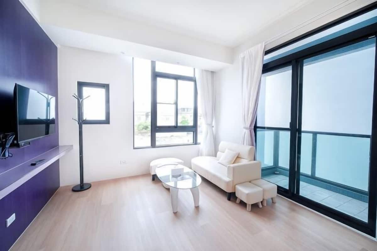 Ein modernes Wohnzimmer mit einer Couch, einem Couchtisch und einem Fernseher. Große Fenster und ein Balkon sind vorhanden.