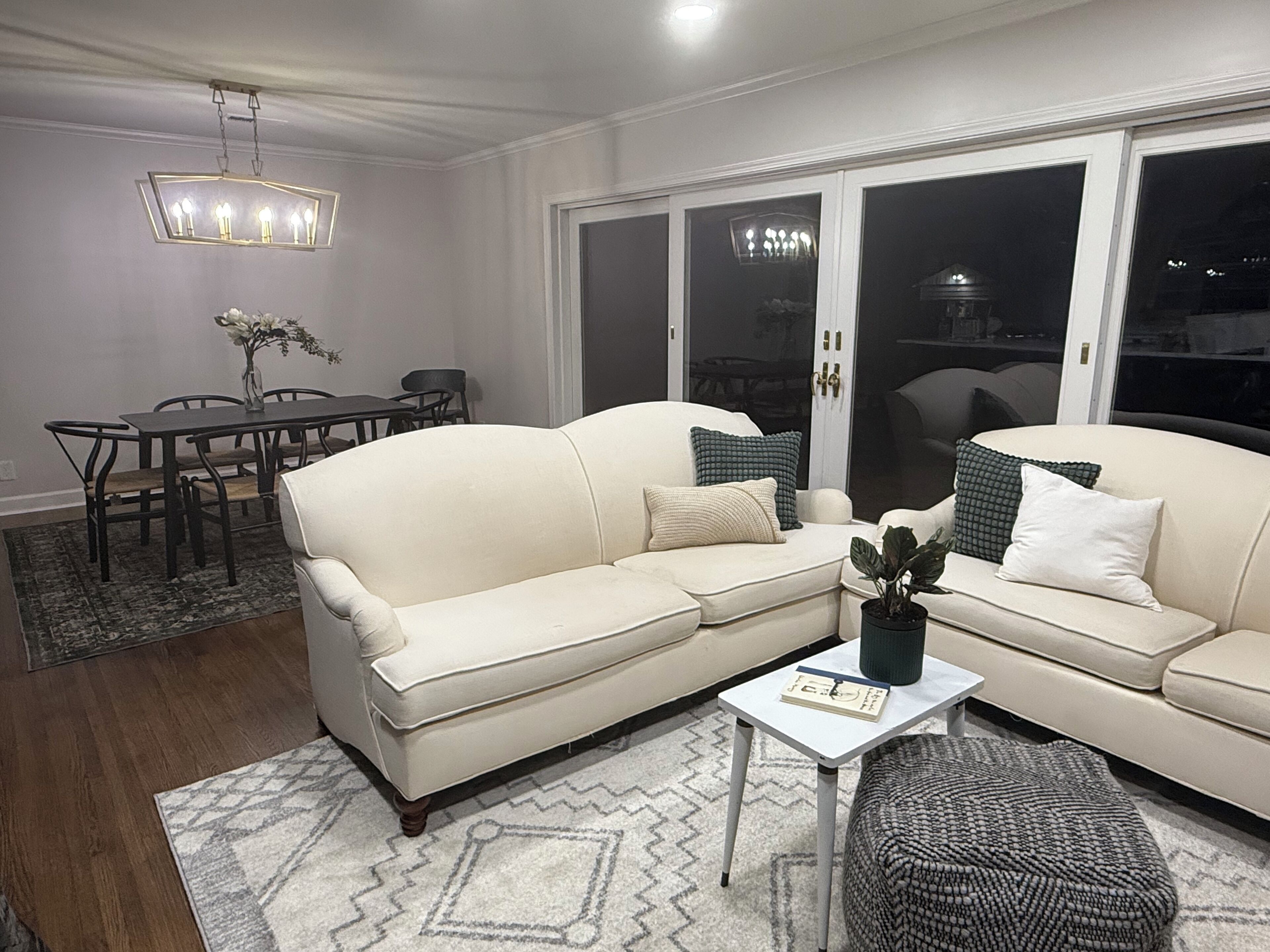A living room with a beige sofa, a coffee table, and a patterned rug. There is a dining table with chairs and a vase with flowers.