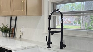 Cucina con piano di lavoro bianco, mobili neri e finestra vista alberi e casa.
