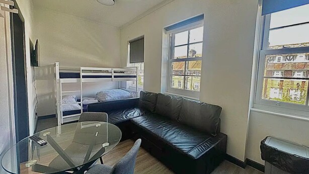 Une pièce de taille modeste, comprenant un lit superposé, un canapé en cuir, une table en verre et une fenêtre donnant sur des immeubles.