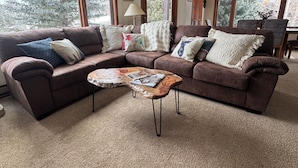 A cozy living room with a brown sectional sofa, a wooden coffee table, and large windows with blinds.
