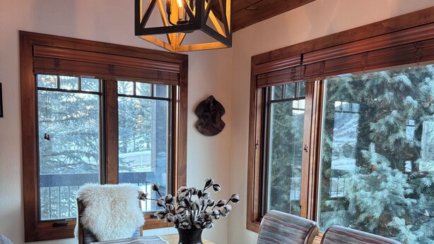 A dining area with a wooden table, chairs, a vase with flowers, a hanging light fixture, and large windows with a view of trees.