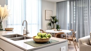 A modern kitchen with a central island, a dining area with a round table and chairs, and large windows with sheer curtains. - Ocean Views at Yves Mermaid Beach (Mermaid Beach)