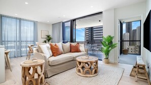 A modern living room with a sofa, two wooden side tables, a balcony with a view of the city, and a large window. - Ocean Views at Yves Mermaid Beach (Mermaid Beach)