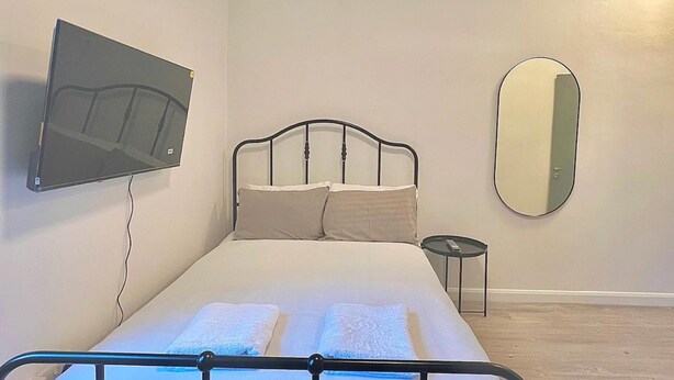 A bedroom with a bed, a TV mounted on the wall, a round mirror, and a small bedside table. - Golders Green Cozy City Studio in London (London)