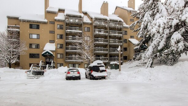 A multi-story building with balconies, a parking area with cars, and snow-covered trees. - Mt Green 2-e06 (Killington)