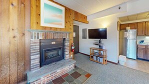 A living room with a fireplace, a television, and a kitchen area with wooden cabinets and a refrigerator. - Mt Green 2-e06 (Killington)