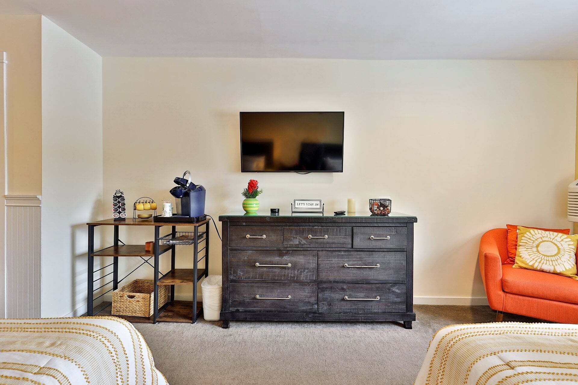 Un salón con un aparador de madera oscura, un televisor montado en la pared, una mesita con utensilios de cocina y un sillón naranja.
