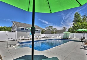 Un patio trasero con piscina, sillas blancas y una sombrilla verde.