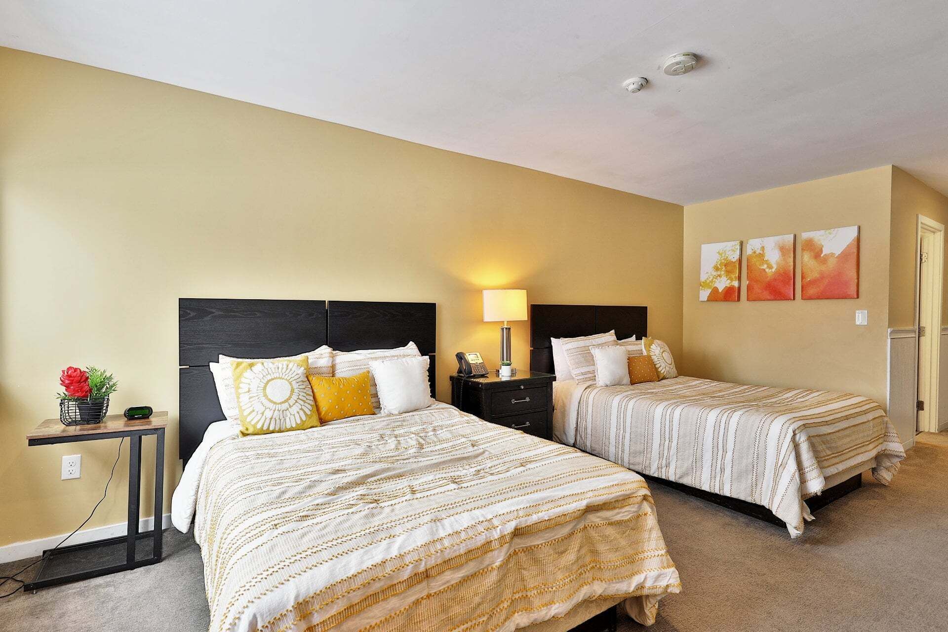 A hotel room with two beds, a bedside table with a lamp, a small table with a flower, and a wall with three paintings.