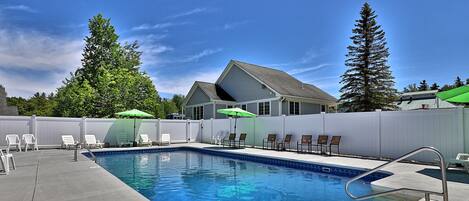 A rectangular swimming pool with a metal ladder, surrounded by white chairs and umbrellas, with a house and trees in the background.