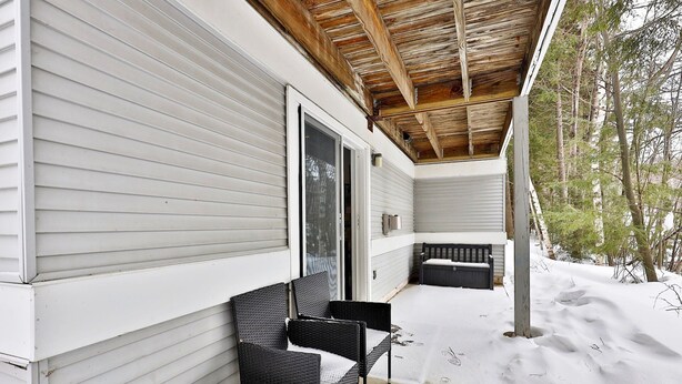 Un portico riparato con due poltrone in vimini nero e un tavolino, soffitto in legno e terreno innevato.