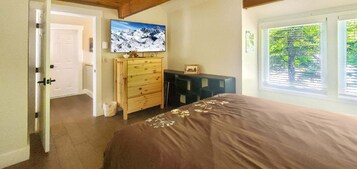 Un dormitorio con cama, cómoda de madera, televisor en la pared y ventana con vistas a árboles.