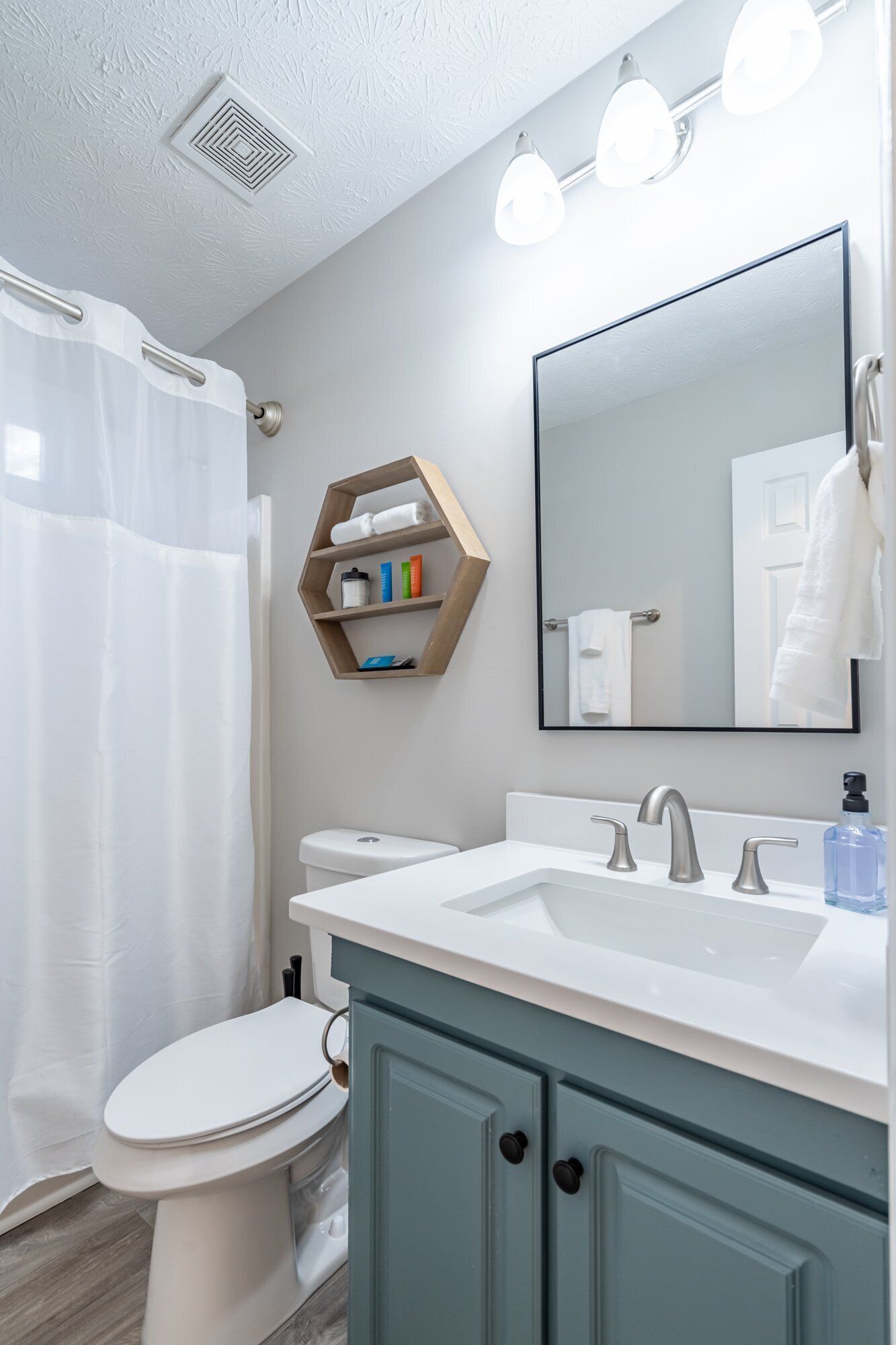 Un baño moderno con inodoro blanco, lavamanos blanco con gabinete azul claro, estante de madera con artículos de aseo personal y un espejo grande.