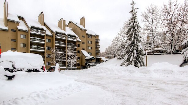 A multi-story building with balconies, surrounded by snow-covered trees and parked cars.