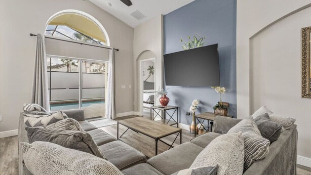 A spacious living room with a large sofa set, a coffee table, and a flat-screen TV mounted on the wall. There is a small table with decorative items and a view of a pool through a glass door. - NEW King Bed Glow In The Dark Kid Cave (Kissimmee)