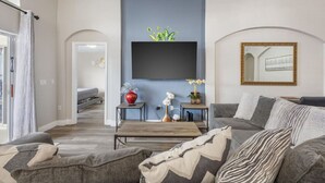 A modern living room with a grey sofa, a coffee table, and a TV mounted on the wall. There is a small table with decorative items and a plant. - NEW King Bed Glow In The Dark Kid Cave (Kissimmee)