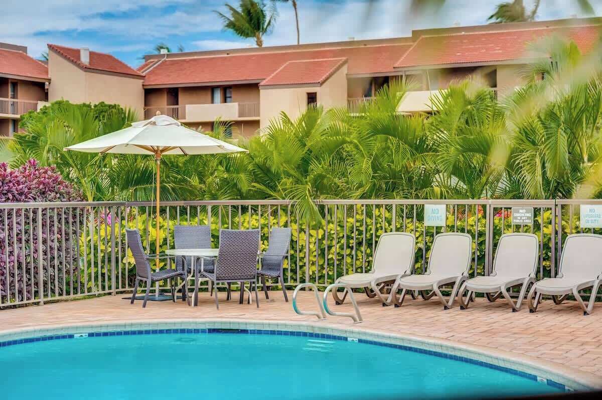 A pool area with lounge chairs and a table, surrounded by a fence and palm trees.